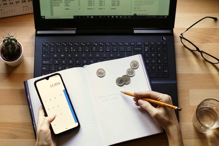 Imagen de una mesa con ordenador abierto. Encima de él una libreta con una calculadora y monedas